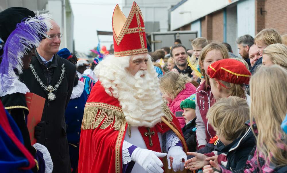 A man with a pointy red hat, frilled red cape, big white beard and long white hair, white gloves, and a cross on his chest, shaking hands with children in a crowd.