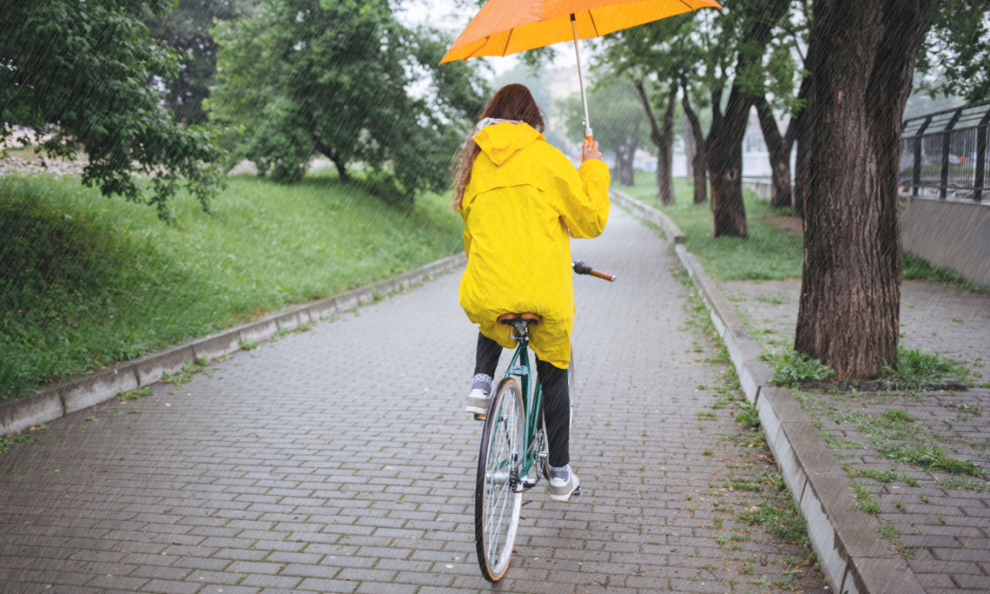 woman wearing yellow rain coat, cycling in rain holding umbrella - safe cycling tips Regina Coeli